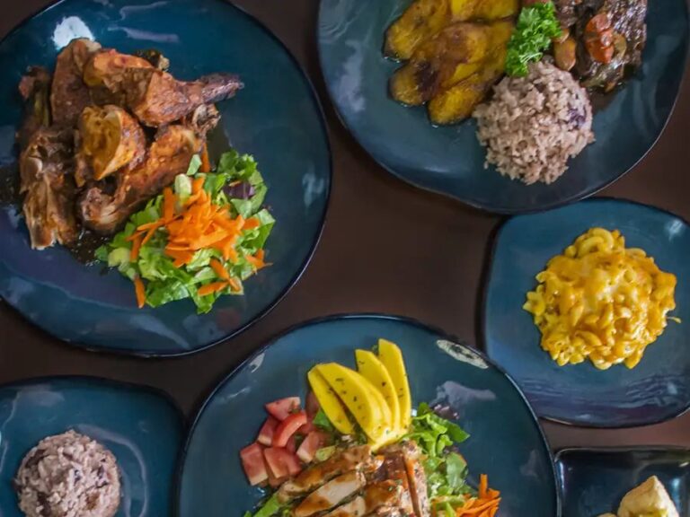 Overhead spread of Jamaican plates with jerk chicken and salad, oxtail with fried plantains and rice and peas, mac and cheese, and a jerk chicken salad